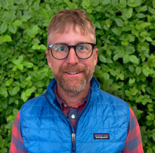 Man with glasses and a blue vest smiling in front of greenery 